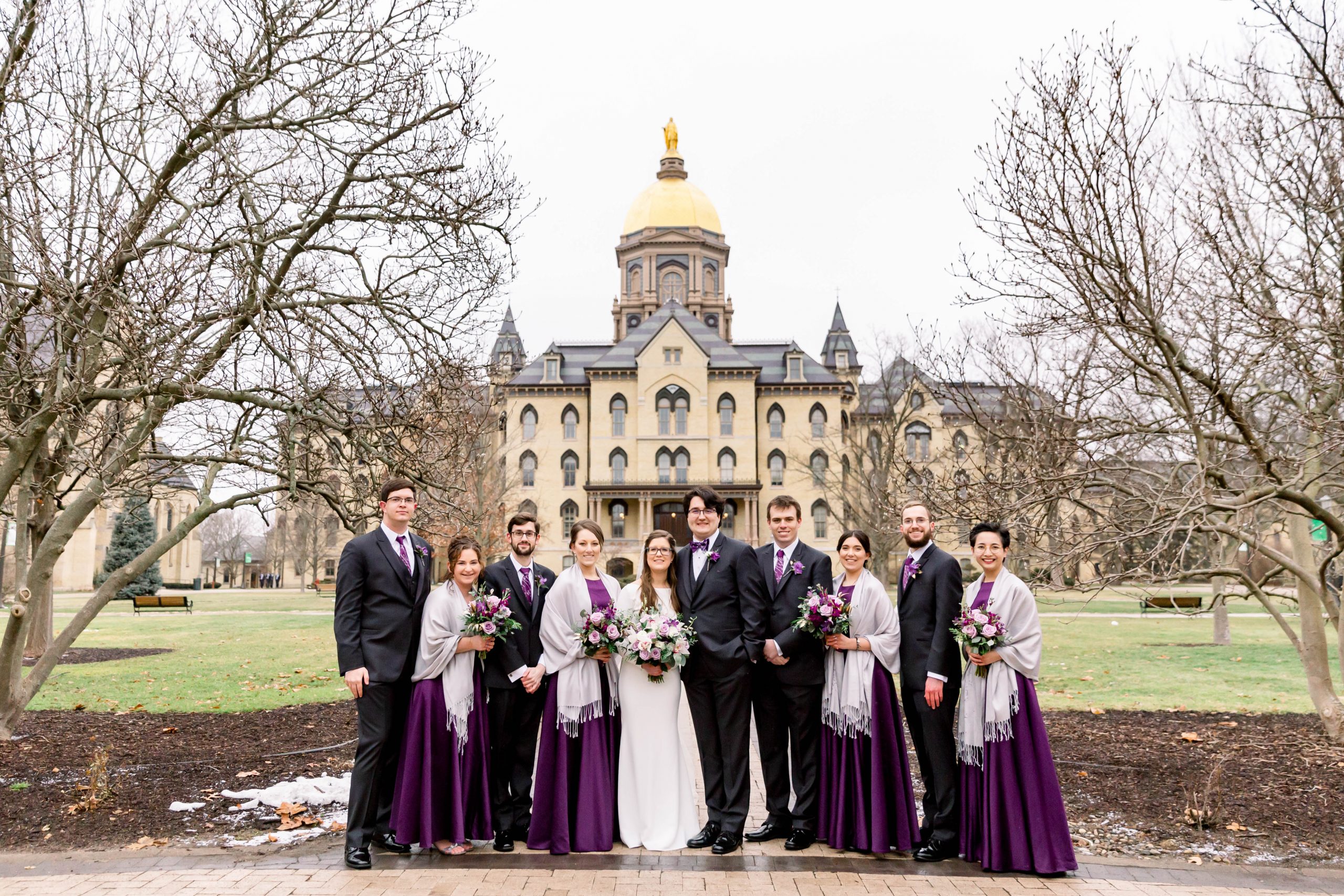 Wedding party standing in front of Notre Dame's main building smiling in long purple dresses and dark gray suits.