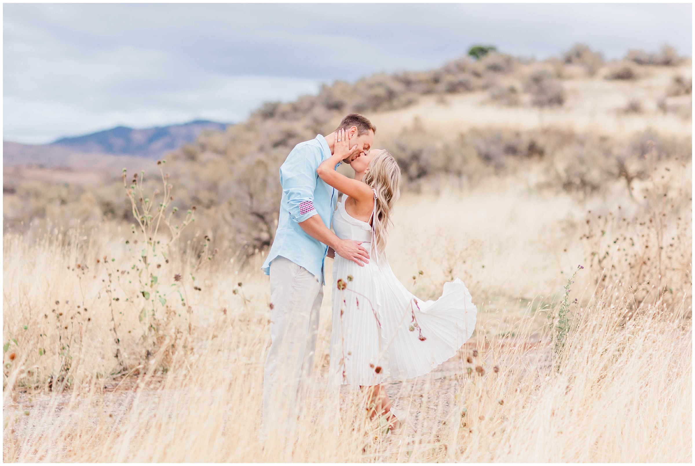 Boise Foothills Engagement photo with a man in a blue shirt kissing a woman in a white flowy dress with light colored sagebrush surrounding them.