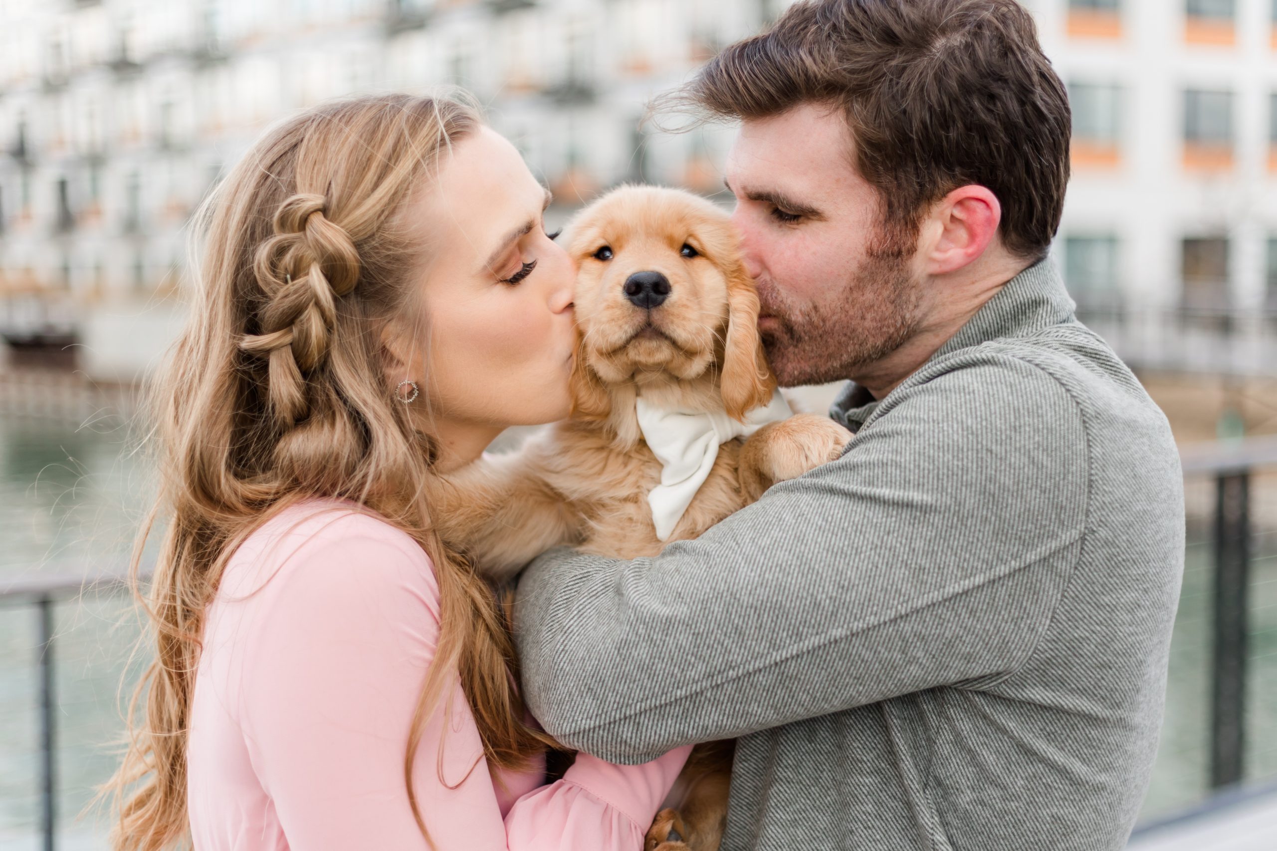 Woman in pink dress and man in green sweater kissing a golden retriever puppy